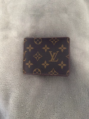 brun plånbok - Snygg brun plånbok från ni vet vart med klassiskt monogrammönster i guld. Plånboken har flera fack för kort och sedlar, och är tillverkad i skinn med lyxig känsla. Perfekt för dig som vill ha en ikonisk accessoar med stil.