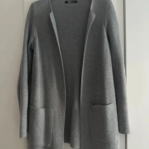 Grå cardigan jacka från Gina Tricot - Snygg grå cardigan jacka från Gina Tricot med lång ärm och två stora fickor framtill. Jackan är stickad och har en öppen front utan knappar eller dragkedja, perfekt för lager på lager. Enkel och stilren design som passar till många outfits