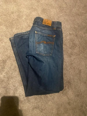 Nudie Jeans Sharp Bengt blå jeans - Snygga Nudie Jeans Sharp Bengt i mörkblå tvätt med klassisk orange söm på bakfickan. Modellen har rak passform och är tillverkad av 100% ekologisk bomull. Jeansens bakficka har en cool slitning för extra edge.
