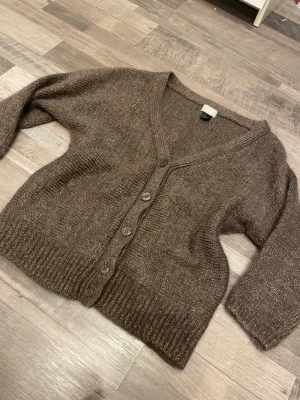 Brun glittrig kofta från H&M - Mysig brun kofta från H&M i storlek M med glittriga inslag i materialet. Koftan har v-ringning, ribbade muddar och fyra knappar framtill. Perfekt för dig som gillar lite extra shimmer i vardagen.