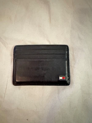 Svart korthållare från Tommy Hilfiger - Snygg svart korthållare i skinn från Tommy Hilfiger med flera kortfack och en liten metallogo framtill. Insidan har ett randigt tygfoder i rött, vitt och blått. Perfekt för dig som vill ha ett stilrent och smidigt alternativ till plånbok.