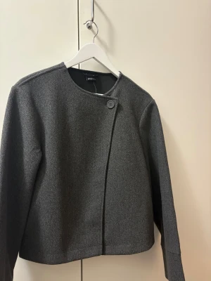 Mörkgrå kavajjacka från Gina Tricot - Stilren mörkgrå kavajjacka från Gina Tricot med rak passform och rund halsringning. Jackan har en snygg asymmetrisk knäppning med en knapp framtill och är helfodrad. Perfekt för dig som gillar minimalistisk och clean stil. Den är heller aldrig använd då etiketten finns kvar.