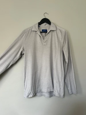 Ljusgrå långärmad pikétröja Eton - Snygg ljusgrå pikétröja från Eton med klassisk krage och lång ärm. Tröjan är tillverkad i mjuk bomull och har en stilren, enkel design utan knappar framtill. Perfekt för dig som gillar en clean och chill look.