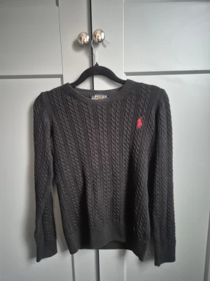 Svart kabelstickad tröja Polo Ralph Lauren - Svart kabelstickad tröja från Polo Ralph Lauren i storlek S. Tröjan har lång ärm, rund hals och den klassiska röda broderade loggan på bröstet. Materialet är mjuk bomull och passformen är normal. Perfekt för dig som gillar stilrena och tidlösa plagg.