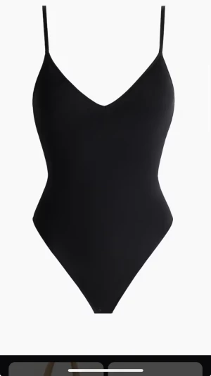 Svart ärmlös bodysuit med v-ringning - Snygg svart bodysuit med smala axelband och v-ringad hals. Modellen är tight och har en clean, minimalistisk look som passar till allt. Materialet är mjukt och stretchigt, vilket gör den bekväm att bära hela dagen. Perfekt att styla med jeans eller kjol.