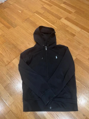 Svart hoodie från Polo Ralph Lauren - Svart hoodie från Polo Ralph Lauren med klassisk vit broderad logga på bröstet. Tröjan har huva med snörning, dragkedja framtill och två fickor. Tillverkad i mjuk bomull, perfekt för chill dagar. Snygg och enkel design som funkar året runt.