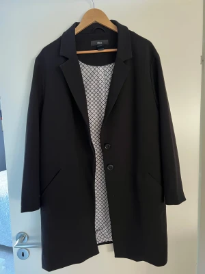 Svart kappa från Ellos, stl 44 - Stilren svart kappa från Ellos i klassisk rak modell med krage och två knappar framtill. Fodret har ett diskret rutmönster. Jackan har långa ärmar och två snedställda fickor. Perfekt för dig som gillar en clean och tidlös look.