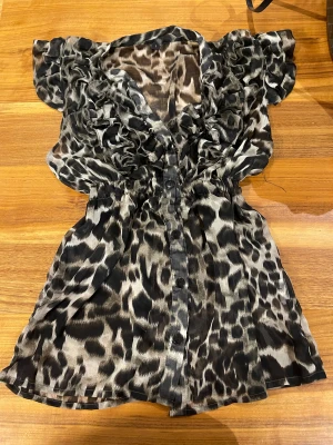 Leopardmönstrad lång blus - Snygg lång blus med leopardmönster och volangdetaljer vid axlar och halsringning. Blusen har knappar framtill och markerad midja. Perfekt för dig som vill sticka ut med en djurisk look.