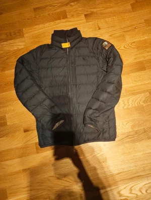 Svart dunjacka från Parajumpers - Svart dunjacka från Parajumpers med quiltad design och coolt patch på ärmen. Jackan har hög krage med gul detalj, dragkedja framtill och långa ärmar. Perfekt för kalla dagar och har en snygg, sportig look.