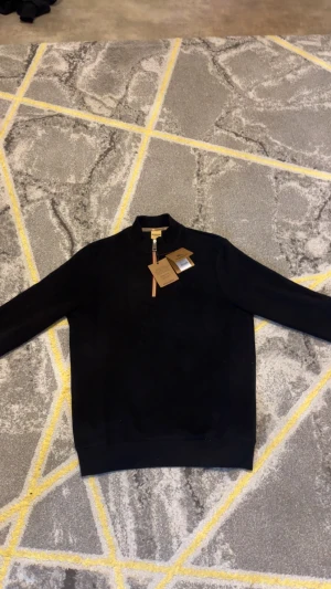 Svart Burberry half zip tröja - Svart långärmad tröja från Burberry med half zip och ribbad krage. Tröjan har ett diskret broderat Burberry-logo på bröstet och dragkedjan har klassiska Burberry-ränder. Tillverkad i ekologisk bomull för en skön och clean känsla.