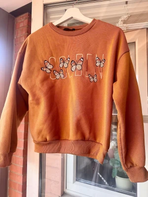 Orange sweatshirt med fjärilar, strl S - Sweatshirt med broderade fjärilar och texten 'GOLDEN' framtill. Tröjan har rund hals, långa ärmar och är tillverkad i mjuk polyester med lite stretch.
