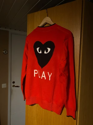 Unik kofta från Comme des Garçons Play - Säljer en unik röd kofta från Comme des Garçons Play med ikoniskt hjärta med ögon på bröstet och stor PLAY-text på ryggen. Koftan har vita knappar, svarta detaljer vid krage och knappslå, samt ribbade muddar. Perfekt för dig som gillar streetstyle och vill sticka ut.