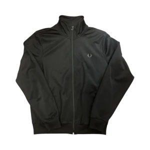 Svart zip hoodie från Fred Perry - Svart zip hoodie från Fred Perry med klassisk ståkrage och hel dragkedja framtill. Tröjan har ribbade muddar vid ärmslut och nederkant samt Fred Perry-loggan broderad på bröstet. Perfekt för en sportig och clean look.