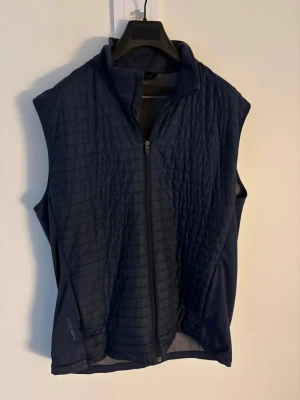 Mörkblå quiltad dunväst (golfväst) Galvin Green - Snygg mörkblå dunväst från Galvin Green med quiltad framsida och hel dragkedja. Västen har en sportig look, hög krage och diskret logga på kragen. Perfekt för lager-på-lager och passar dig som gillar stilren design. Storlek: L passar M Nypris: 1200kr