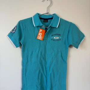 Turkos pikétröja - 134/140 - Turkos pikétröja med vit kant på krage och ärmslut, broderad text på bröstet och siffror på ärmarna. Klassisk piké med knappar framtill.