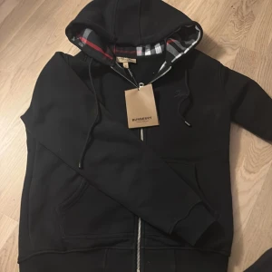 Svart hoodie från Burberry med dragkedja - Svart hoodie från Burberry i bomull med klassiskt rutigt foder i huvan, dragkedja framtill och broderad logga på bröstet. Snygg och stilren med ribbade muddar och fickor på sidorna. Perfekt för dig som vill ha en exklusiv streetstyle-look.