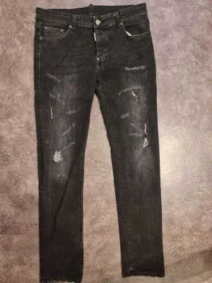 Svarta jeans från Dsquared2, storlek 50 - Svarta jeans från Dsquared2 med slitna detaljer och snyggt tvättad look. Klassisk femficksmodell med knappgylf och svart läderpatch bak i midjan. Jeansen har en rak passform och är tillverkade i jeanstyg med coola slitningar framtill.