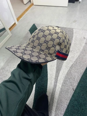 Gucci GG supreme keps äkta - Säljer en snygg beige keps från Gucci med klassiskt GG-mönster i mörkblått. Kepsen har en justerbar rem i mörkblått skinn och en röd-blå detalj på sidan. Tillverkad i Italien med exklusiv känsla och cool design, perfekt för dig som gillar streetstyle. Har inget kvitto priset kan diskuteras 