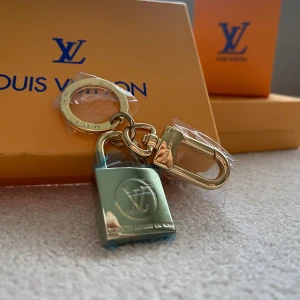 Vuitton guld nyckelring med hänglås - Exklusiv nyckelring från  Vuitton i glänsande guldmetall med ikoniskt -hänglås. Ringen har ingraverad logga och robust karbinhake, perfekt som accessoar till väskan eller nycklarna. Lyxig och stilren detalj för dig som gillar designer.