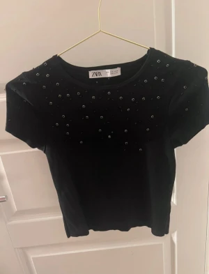 Svart croppad topp med pärlor från Zara - Svart croppad topp från Zara med korta ärmar och rund hals. Toppen är dekorerad med små svarta pärlor framtill och har en tight passform som framhäver midjan. Perfekt för dig som gillar stilrena detaljer och vill sticka ut lite extra. Endast använt en gång