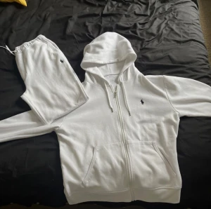 Vit Polo Ralph Lauren dress   - Vit hoodie från Polo Ralph Lauren med dragkedja och huva. Klassisk broderad logga i svart på bröstet, snörning vid huvan och två fickor framtill. Tillverkad i mjuk bomullsmix som är skön att ha på sig. Perfekt för chill dagar eller när du vill ha en clean look.