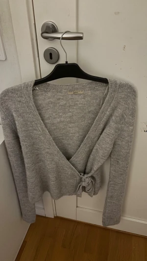 Grå omlottkofta från Gina Tricot - Mysig grå stickad omlottkofta från Gina Tricot med lång ärm och knyt i sidan. Perfekt för lager-på-lager och har en snygg v-ringad design. Koftan är i mjukt material och passar dig som gillar enkel och stilren look.