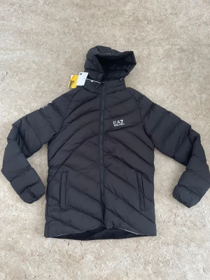 Svart pufferjacka från EA7 Emporio Armani - Snygg svart pufferjacka från EA7 Emporio Armani med huva och dragkedja framtill. Jackan har diskret logga på bröstet och huvan, samt två sidofickor. Perfekt för kalla dagar med sin vadderade design och stilrena look.