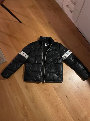 Svart pufferjacka från Calvin Klein - Svart pufferjacka från Calvin Klein Jeans med vita CK-loggor på ärmarna och dragkedja framtill. Jackan har hög krage, två fickor med dragkedja och quiltad design. Perfekt för kalla dagar. Den är för liten för mig så säljer den
