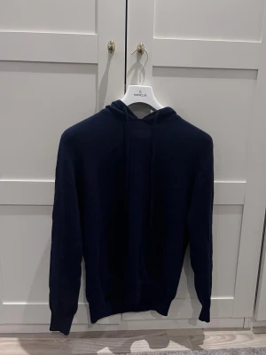 Mörkblå hoodie i kashmir  - Snygg mörkblå hoodie i mjuk kashmir. Tröjan har huva med snörning och långa ärmar, perfekt för dig som gillar stilren och lyxig känsla. Passformen är normal och materialet är riktigt skönt mot huden. En klassisk hoodie som funkar året runt.