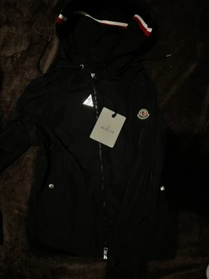 Svart vindjacka från Moncler - Svart vindjacka från Moncler med huva och snörning, dekorerad med röd och vit rand på insidan av huvan. Jackan har dragkedja framtill, silverfärgade knappar på fickorna och Moncler-logga på bröstet. Perfekt för dig som gillar stilren.