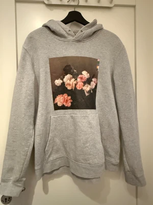 Grå hoodie med blommigt tryck - Säljer en grå hoodie i storlek M med ett stort blommigt tryck på bröstet i rosa och beige toner. Tröjan har huva med snörning och en klassisk magficka. Perfekt för dig som gillar konstnärliga detaljer och chill stil.