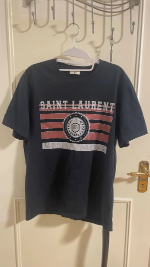 Svart Saint Laurent t-shirt med tryck - Cool svart t-shirt från Saint Laurent med vit och röd grafisk print på bröstet och logga. Klassisk rund hals och korta ärmar. Materialet är mjuk bomull som känns skönt mot huden. Perfekt för dig som gillar streetstyle och designer.
