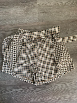 Rutig shorts med bälte från H&M  - Snygga rutig shorts från H&M i beige, svart och vit med tillhörande bälte. Modellen är högmidjad och har en loose passform. Materialet är en mix av polyester, viskos och elastan som känns mjukt och skönt. Perfekt för dig som gillar trendiga och bekväma plagg.