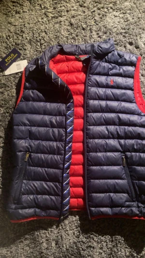Mörkblå och röd dunväst från Polo Ralph Lauren - Snygg dunväst från Polo Ralph Lauren i mörkblått med röda detaljer och insida. Västen har quiltad design, dragkedja framtill och två fickor med dragkedja. Klassisk Polo-logga broderad på bröstet. Perfekt för lager-på-lager och chill stil. Pris kan diskuteras 