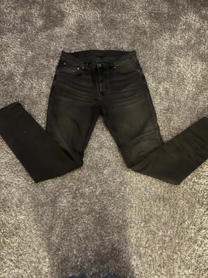 Mörkgråa jeans från Nudie - Säljer ett par mörkgråa  jeans från Nudie med snyggt slitna detaljer och klassiska bakfickor med vågigt broderi. Jeansen har fem fickor, dragkedja och är tillverkade i stretchig bomullsdenim. Nypris- 1300kr/mitt pris-350kr!
