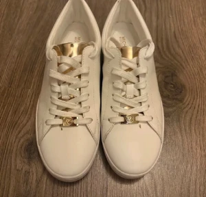 Vita sneakers från Michael Kors - Snygga vita sneakers från Michael Kors med gulddetaljer på plösen och snörningen. Skorna har rund tå, platt sula och är tillverkade i skinn. Diskret MK-logga på sidan och snörning framtill. Perfekta för dig som gillar stilrena och lyxiga sneakers.
