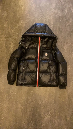 Svart Moncler Montbeliard dunjacka med huva - Svart glansig dunjacka från Moncler med stor huva och klassisk logga på bröstet. Jackan har en röd-vit-blå rand längs dragkedjan, två fickor med dragkedja och quiltad design. Perfekt för kalla dagar och riktigt snygg streetstil.