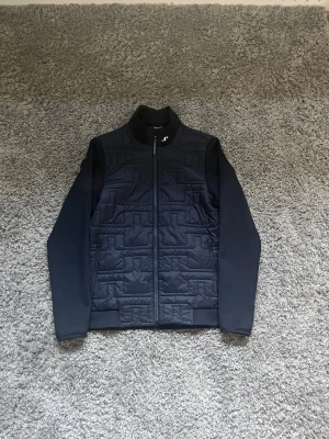 Mörkblå quiltad jacka J.Lindeberg - Snygg mörkblå jacka från J.Lindeberg med quiltad framsida och släta ärmar. Jackan har dragkedja hela vägen, hög krage och diskret logga på bröstet. Materialet är polyester och jackan har en modern, normal passform. Perfekt för dig som gillar stilren design.