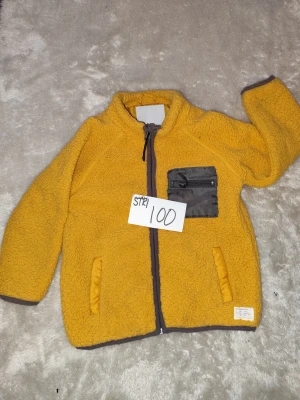 teddyjacka - Gul teddyjacka.    Jackan har en cool bröstficka med dragkedja i mörkare tyg och två sidofickor. Mjuk och fluffig känsla, perfekt för kyliga dagar. Långärmad och med kontrastkant vid ärmslut och nederkant.... Användes som tjock tröja särskilt i bilen ist för jacka . 