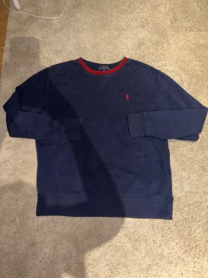Mörkblå sweatshirt från Polo Ralph Lauren - Snygg mörkblå sweatshirt från Polo Ralph Lauren med röd broderad logga på bröstet och röd/blå rand vid halsen. Tröjan har långärmad passform och är gjord i mjuk bomull. Perfekt för chill dagar och enkel att matcha med jeans eller joggers.