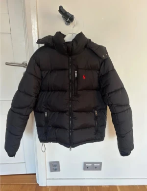 Svart pufferjacka från Polo Ralph Lauren - Svart pufferjacka från Polo Ralph Lauren med röd logga på bröstet. Jackan har huva, flera dragkedjeförsedda fickor och quiltad design. Perfekt för kalla dagar med sin tjocka och fluffiga look. Snygg och stilren modell som håller värmen.