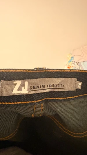 Blå wide jeans från ZJ Denim Identity - Snygga blå jeans från ZJ Denim Identity i storlek 46. Modellen har raka och breda ben, klassiska fem fickor och kontrasterande gula sömmar. Materialet är 99% bomull och 1% spandex för lite stretch. Benlängd 84 cm.
