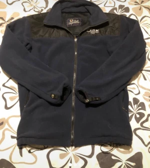 Mörkblå fleecejacka Peak Performance - Snygg mörkblå fleecejacka från Peak Performance med svarta axelpartier och broderad logga. Jackan har hel dragkedja, höga fickor och justerbara ärmslut med knapp. Perfekt för kyliga dagar och outdoor-häng.