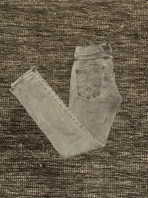 Replay grå jeans - Säljer ett par grå jeans från Replay med klassisk femficksdesign och diskreta sömmar på bakfickorna. Jeansen har låg midja och har ett stretchigt jeanstyg för en tight passform. Perfekta för dig som gillar en clean och modern look.