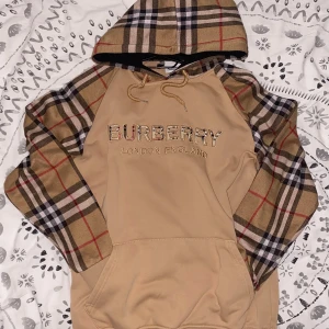 Sälsynt burberry hoodie - Burberry hoodie i beige med klassiska rutiga ärmar och huva. Stor logga broderad på bröstet. Tröjan har snörning vid huvan och en stor magficka. Tillverkad i 100% bomull för skön känsla. Perfekt för dig som gillar lyxiga streetwear-vibbar. Passar för er runt 165-175 Har inga fel inga defekter har nästan aldrig använts och priset kan prutas ner vid snabb affär även om den inte är så saltig. SKRIV VID INTRESSE ANNARS SÄLGS DEN TILL NÅGON ANNAN:))