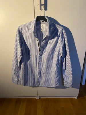 Blårandig skjorta från Lacoste - Snygg blårandig skjorta från Lacoste med klassisk krage och slim fit-passform. Skjortan har långa ärmar, knappar framtill och den ikoniska krokodilloggan på bröstet. Tillverkad i mjuk bomull som känns skön mot huden. Perfekt för dig som gillar stilrena och fräscha plagg.