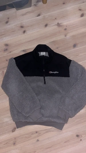 Grå champion fleece jacka  - Säljer en grå och svart fleece-tröja från Champion i storlek medium. Tröjan har en mysig teddystruktur, svart panel upptill med broderad Champion-logga och praktisk half zip framtill. Perfekt för kyliga dagar och chill outfits.
