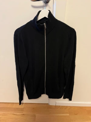 Svart kofta med dragkedja från Jack and Jones  - Svart kofta med hög krage och hel dragkedja framtill. Tröjan har långa ärmar och två fickor på sidorna. Enkel och stilren design som passar till många olika outfits. Materialet känns mjukt och bekvämt mot huden.