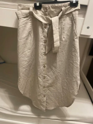 Beige linnelångkjol med knappar och bälte - Stilren beige långkjol i linne med knappar framtill och tillhörande bälte i midjan. Kjolen har fickor på sidorna och en lätt rundad nederkant. Perfekt för dig som gillar en avslappnad men ändå chic look.