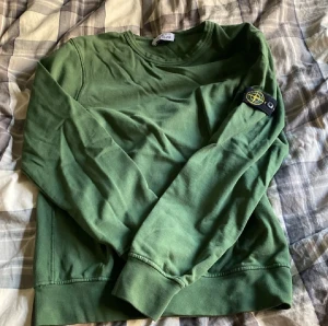 Grön långärmad tröja Stone Island - Säljer en grön långärmad tröja från Stone Island Junior med klassisk rund hals och ikonisk patch på vänster ärm. Tröjan är i mjuk bomull och har ribbade muddar vid ärmslut och nederkant. Perfekt för dig som gillar streetwear och vill ha något stilrent. Passar 12-13 åringar